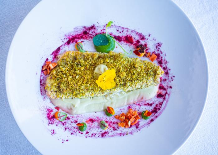Piatto elegante con crosta di pistacchio, crema e fiori colorati.
