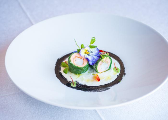 Piatto elegante con rotolini verdi, crema bianca, fiori colorati e dettagli raffinati.